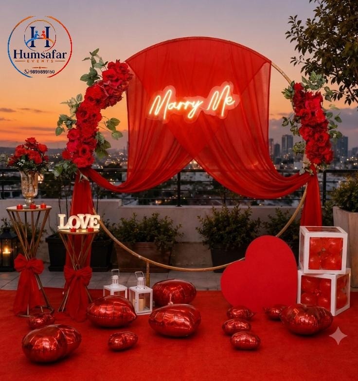 ​Gold foil balloons spell "BE MY LOVE" beside a bed adorned with a red rose petal heart, complemented by a ceiling and floor filled with metallic rose gold, sage green, and white balloons.